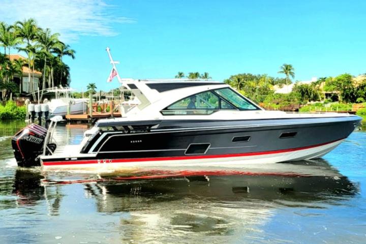A motorboat with black hull and red accents on a calm waterway, surrounded by greenery and blue sky.